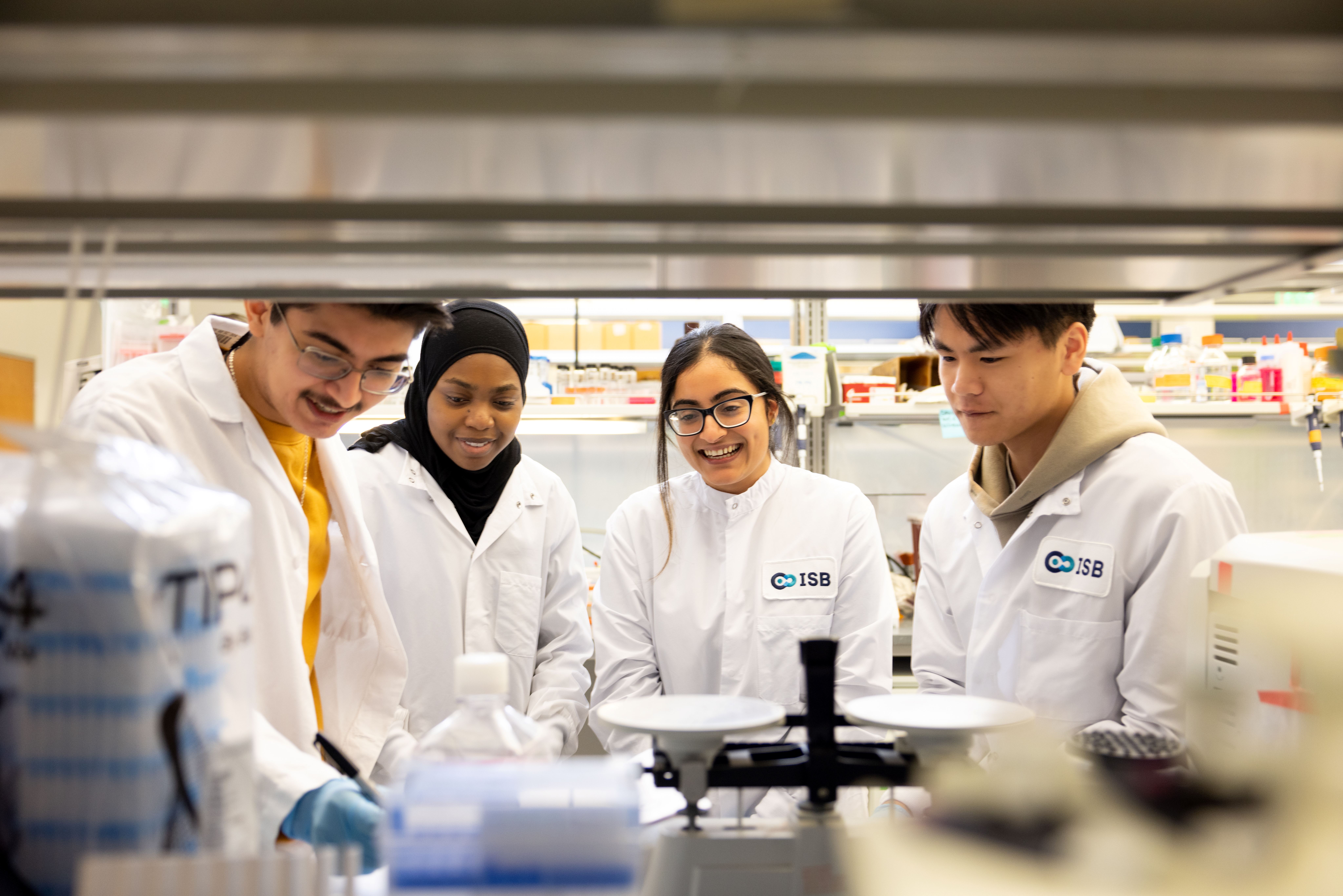Luis, Rehema, Alisha, & Brian in a science lab wearing white lab coats