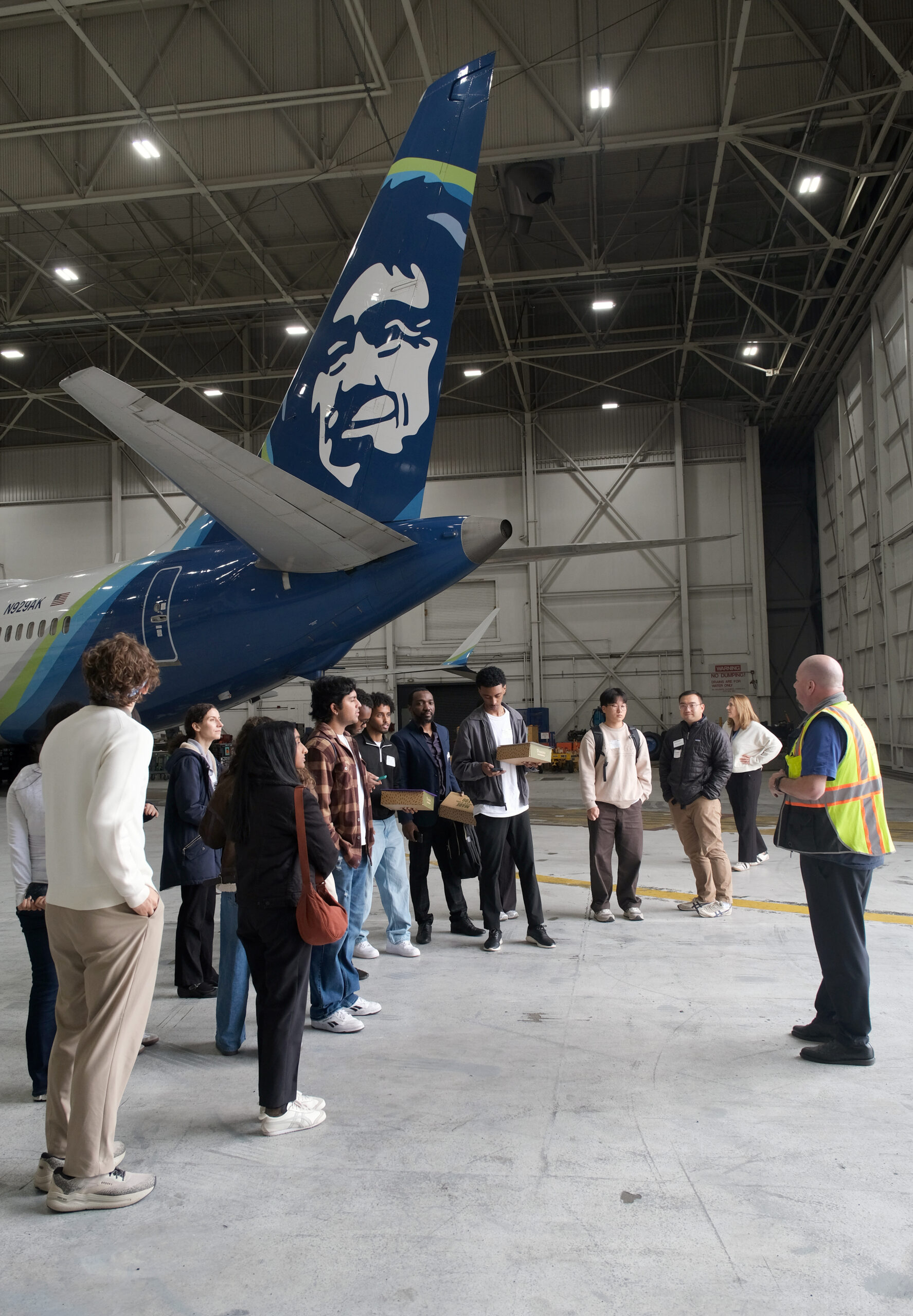 A group of Scholars standing in an Alaska Airlines hangar while a man in a yellow safety vest speaks to them.