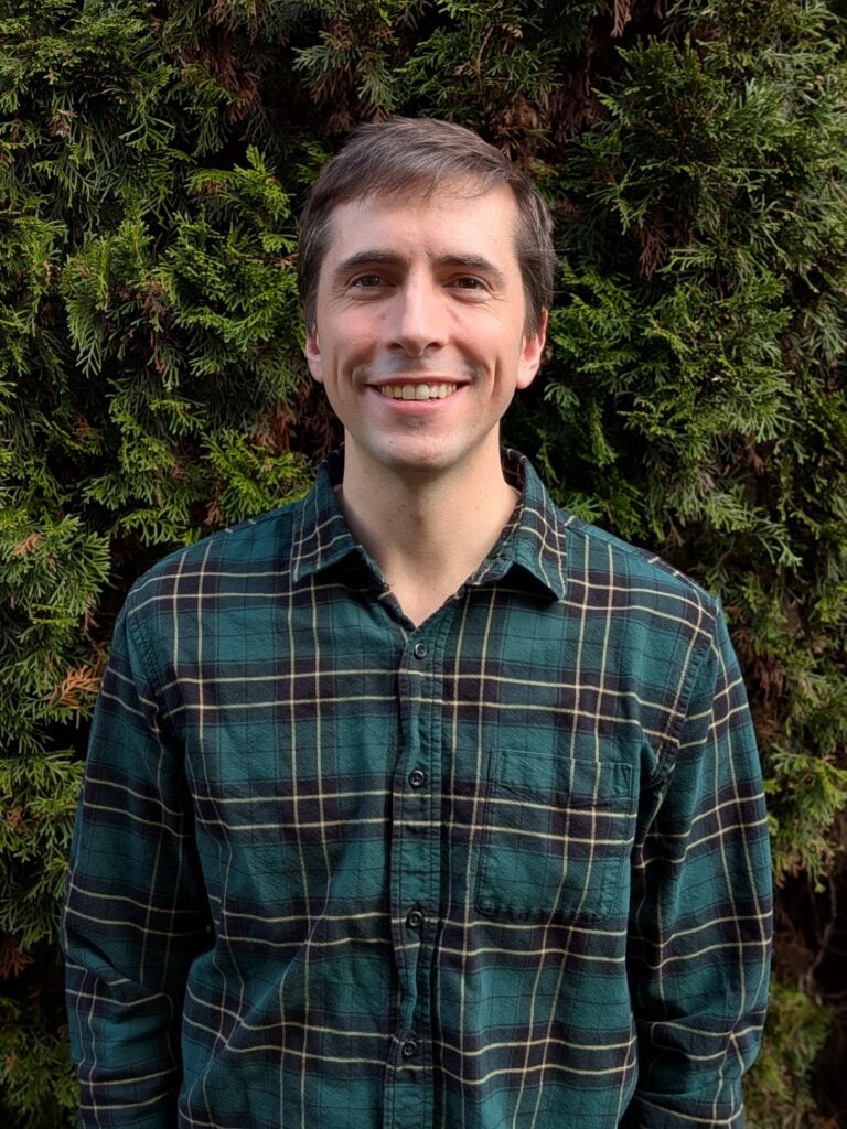 photo of a white male in a green and blue flannel button up in front of a green bush